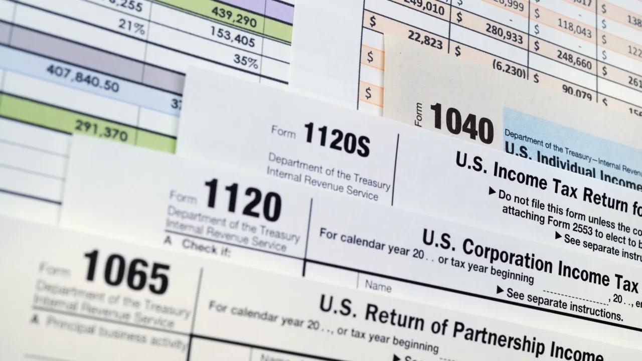 IRS forms 1065, 1120, 1120S and 1040 fanned out in overlapping layers across a printed spreadsheet.