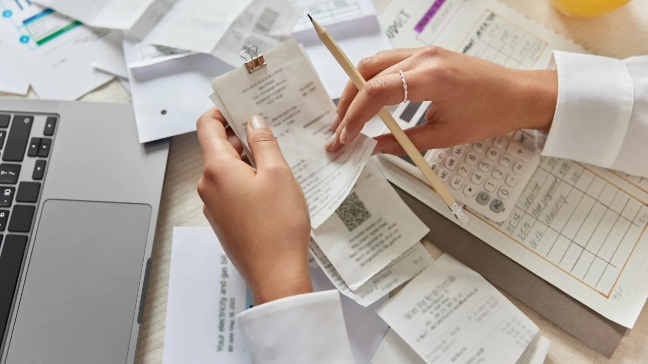 A pair of hands sorting a pile of paper receipts on a desk crowded with a laptop, a calculator and a hand-ruled ledger.